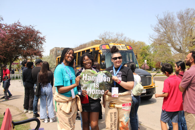 Three people smiling, holding a "Hamilton is Home" sign in front of a school bus. Around them, a diverse crowd stands under a clear sky, giving a sense of community and belonging.