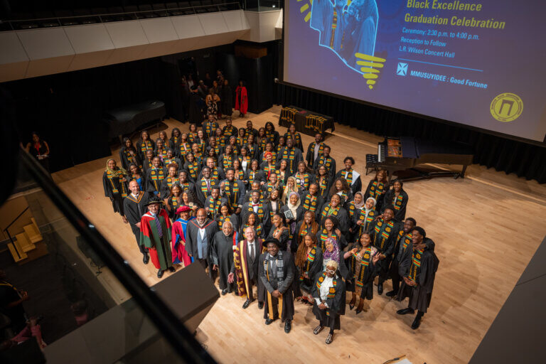 A group of graduates in black gowns with colorful stoles pose on a stage, with faculty in academic regalia in the front row during Black Excellence Graduation Celebration.