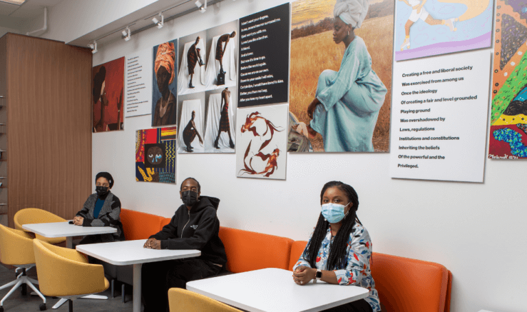 Faith Ogunkoya and two students sit at tables in a room adorned with vibrant artworks on the wall.