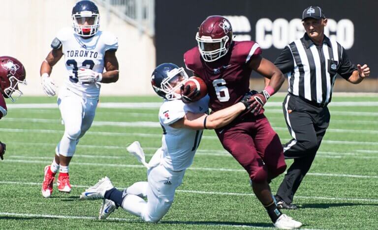 Football game with a player in a maroon uniform running with the ball, being tackled by an opposing player in white.