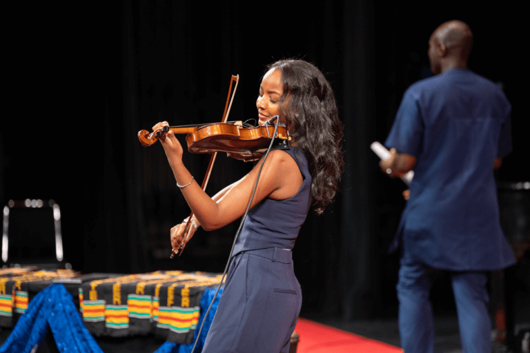 A woman plays the violin on stage while a man stands in the background holding a rolled paper.
