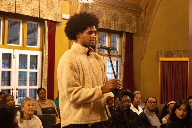 A person speaks into a microphone in front of an audience in a warmly lit room with large windows and decorative curtains.