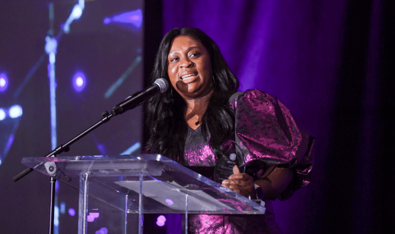 Faith Ogunkoya speaking at a podium with a microphone, holding an award, against a purple-lit background.