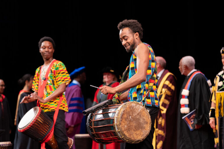 Two musicians in colorful traditional attire play drums on stage, while people in academic regalia stand in the background.