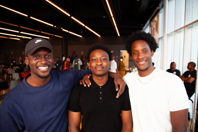 Three friends smiling warmly in a modern, well-lit indoor setting. They appear relaxed and happy, with two of them arm in arm, conveying camaraderie.