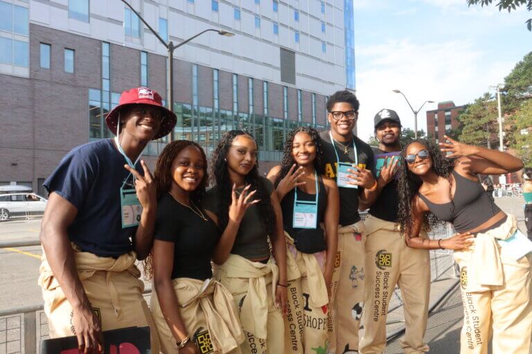A group of seven young people smiling and posing outside in front of a modern building.