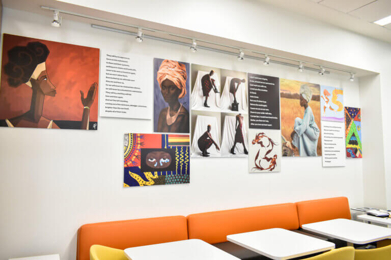 A wall with various artworks, photographs, and poetic texts displayed above an orange bench and white tables.