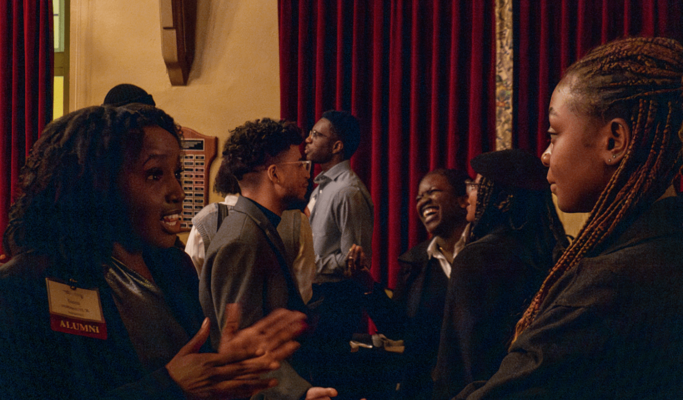 A lively gathering of diverse alumni engaging in animated conversations indoors. The atmosphere is warm, with people smiling and laughing against a backdrop of red curtains.