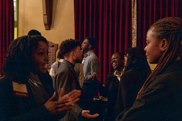 A lively gathering of diverse alumni engaging in animated conversations indoors. The atmosphere is warm, with people smiling and laughing against a backdrop of red curtains.
