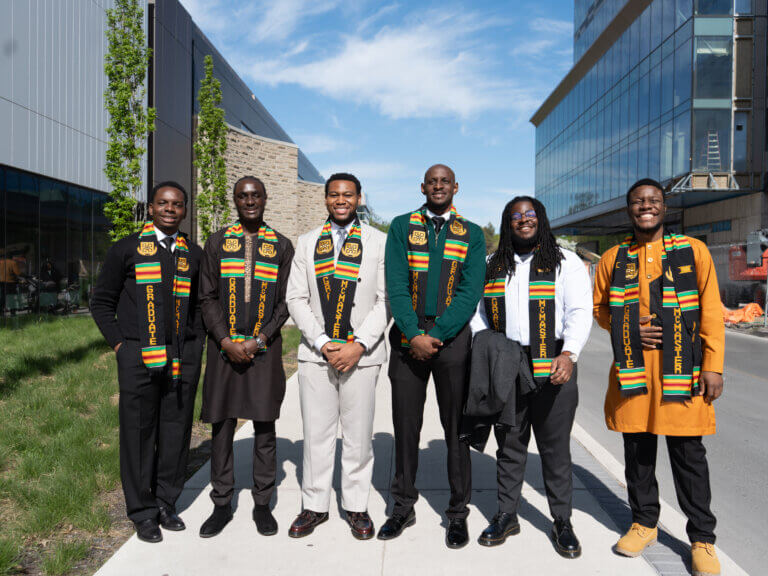 Six people in colorful stoles pose outdoors, smiling under a clear sky. Modern buildings and greenery surround them, conveying a celebratory mood.