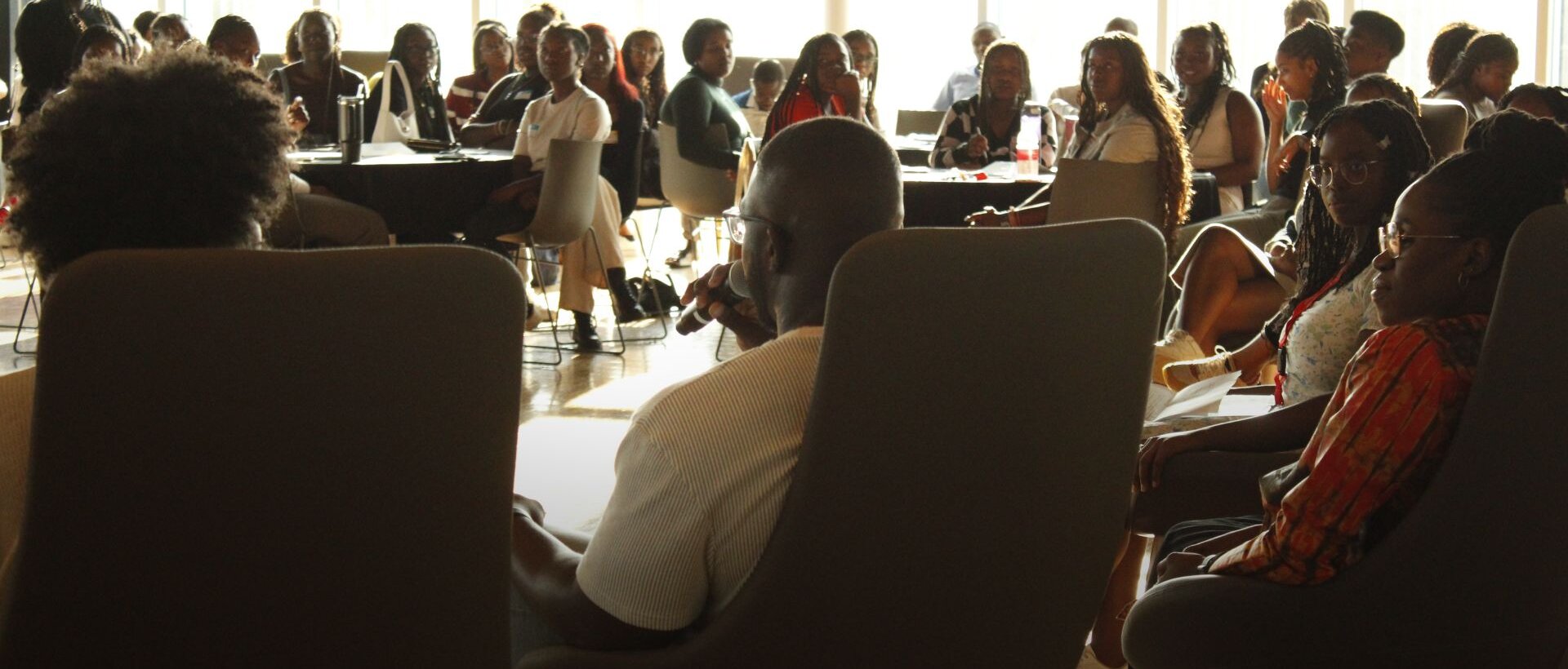 A diverse group of people seated in a well-lit conference room, attentively listening to a speaker. The atmosphere is focused and engaged.