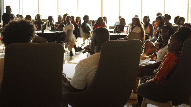 A diverse group of people seated in a well-lit conference room, attentively listening to a speaker. The atmosphere is focused and engaged.