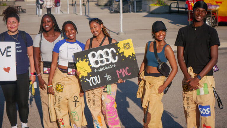 A group of six smiling people stand outdoors holding a colorful sign saying