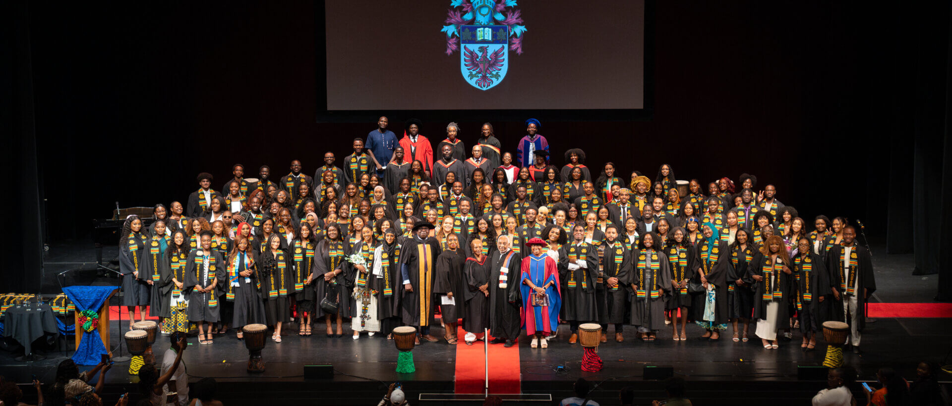 A large group of students in graduation gowns and stoles stand on stage, smiling. A crest is projected above them. The atmosphere is celebratory.