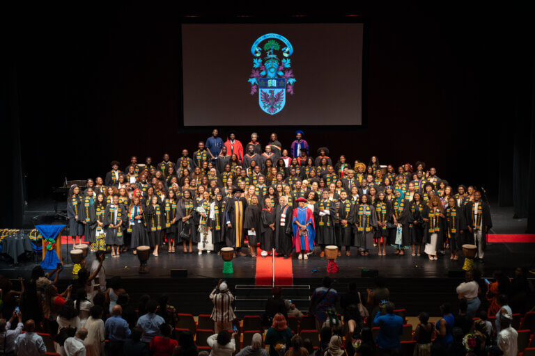 A large group of students in graduation gowns and stoles stand on stage, smiling. A crest is projected above them. The atmosphere is celebratory.