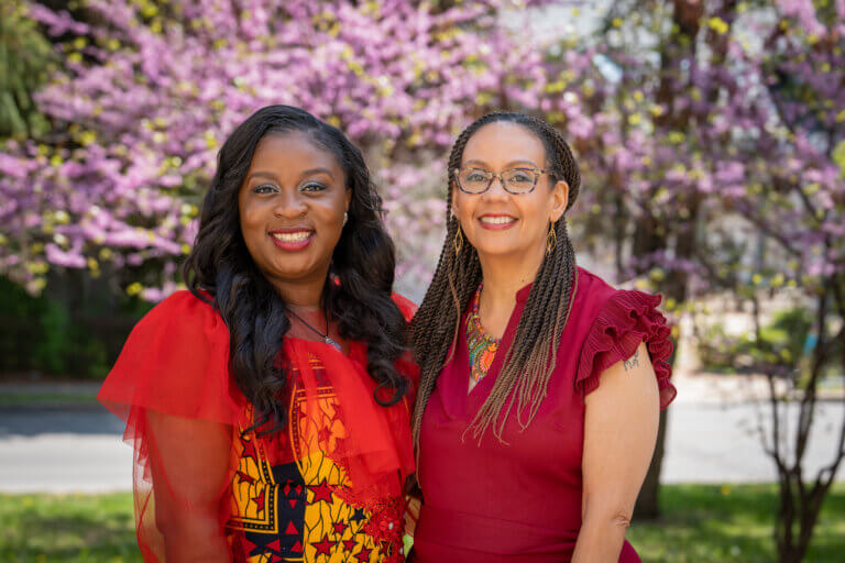 Two smiling women in colorful attire stand outdoors with a backdrop of blooming purple trees. The scene is bright and joyful, reflecting a sense of warmth.