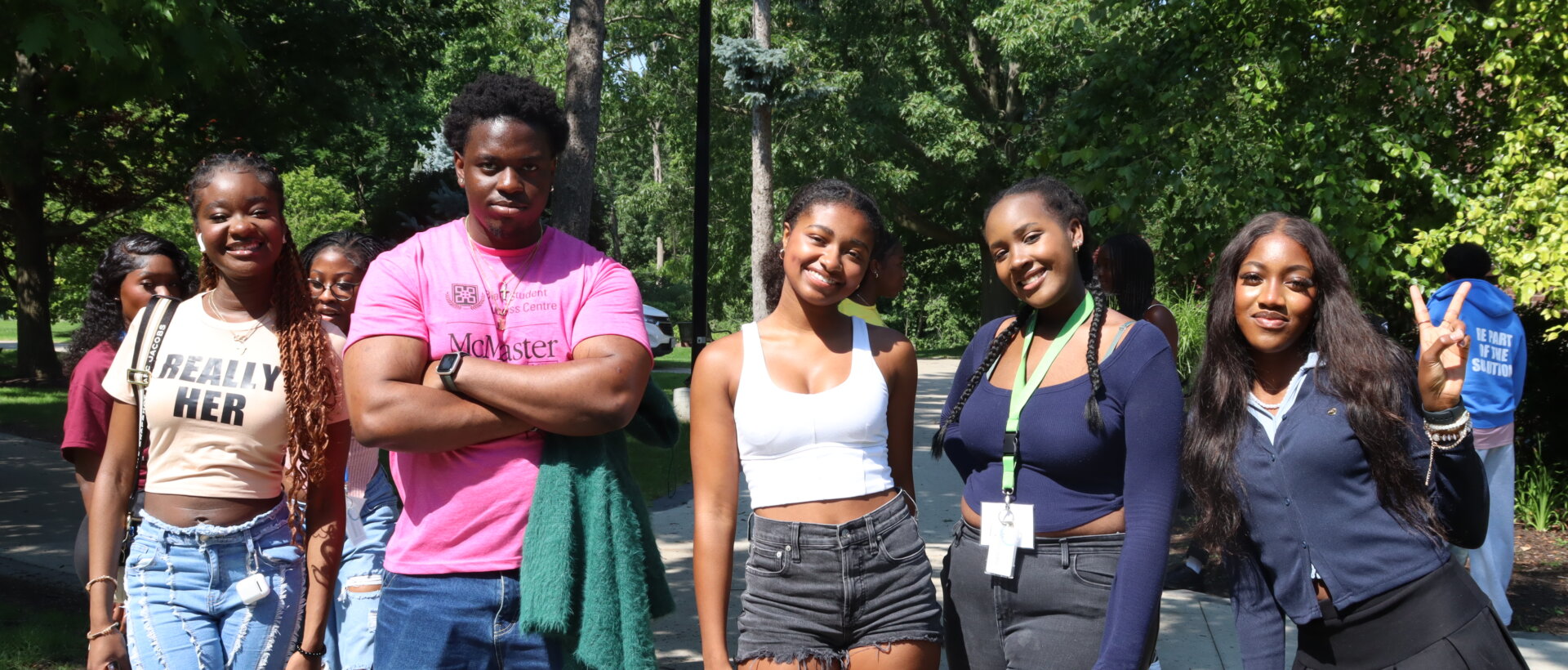 A group of five young adults smiling outdoors on a sunny day. They're casually dressed, standing on a path surrounded by lush green trees.