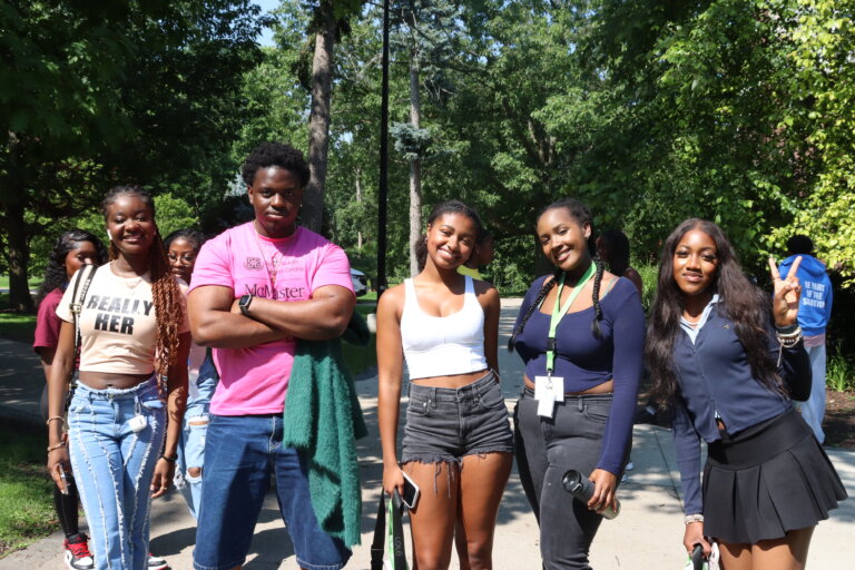 A group of five young adults smiling outdoors on a sunny day. They