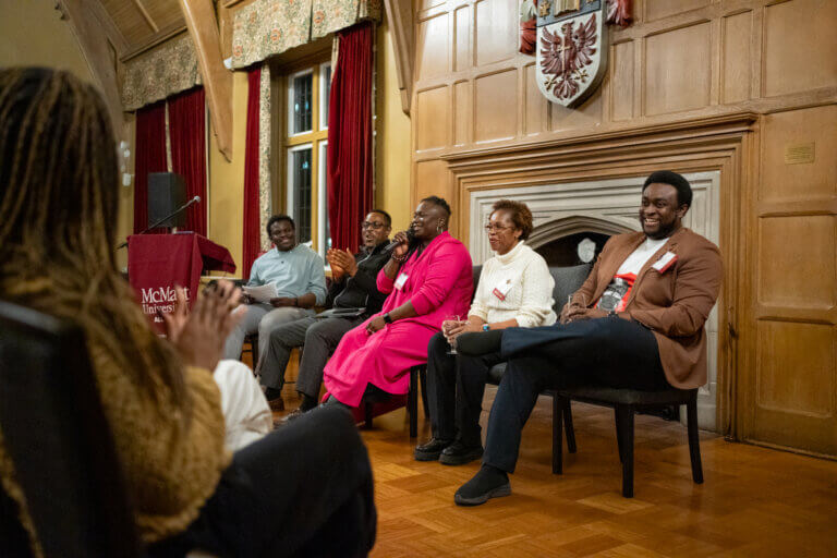 A panel of five people sit in a warmly lit room with wooden walls and red curtains, engaging in a lively discussion. Audience member clapping in foreground.
