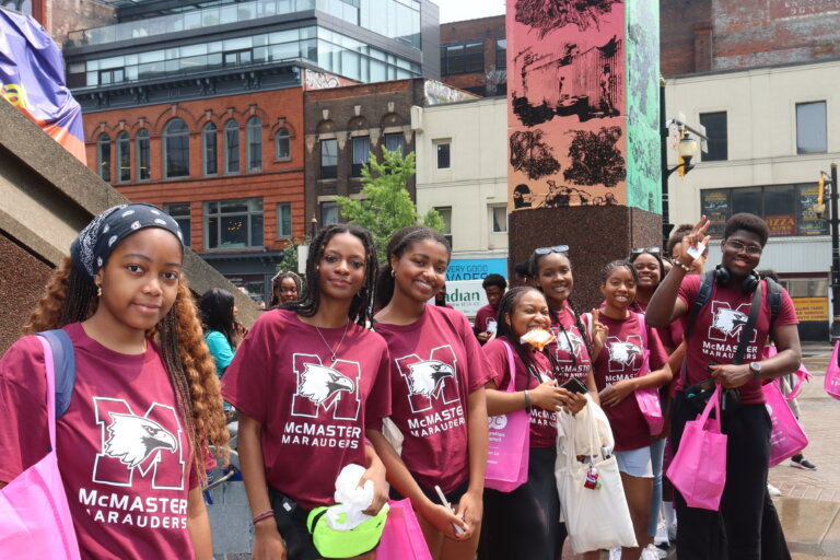 A group of smiling students in maroon McMaster Marauders shirts hold pink bags, standing outdoors in a city square with brick buildings in the background.