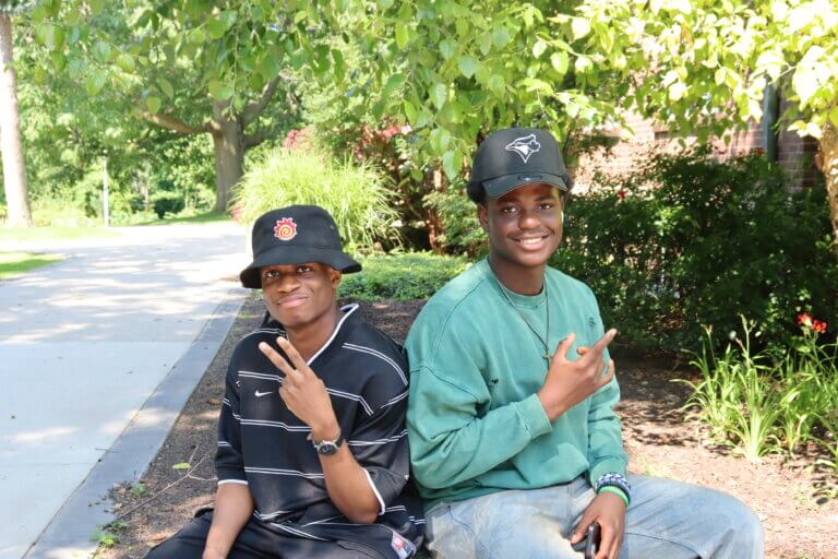 Two people sit on a bench in a sunny park, smiling and making peace signs. They wear casual clothes and caps, surrounded by greenery and flowers.
