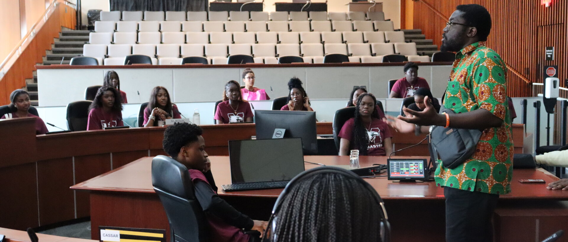 A man in a colorful shirt speaks to attentive students in a lecture hall. They are seated in a semi-circle with laptops open, creating an engaged and educational atmosphere.