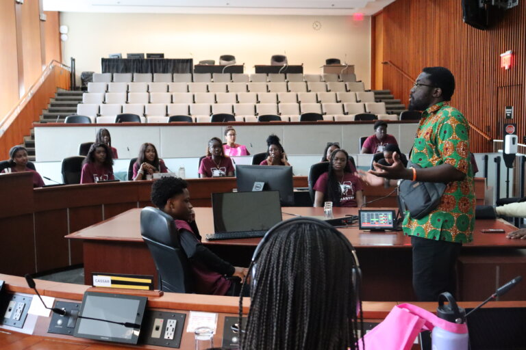 A man in a colorful shirt speaks to attentive students in a lecture hall. They are seated in a semi-circle with laptops open, creating an engaged and educational atmosphere.
