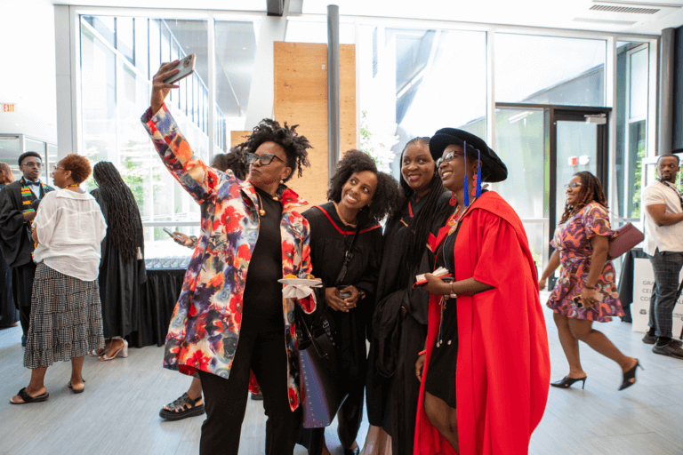 A joyous group of four people, dressed in graduation attire, gather for a selfie. One holds the phone aloft while others smile warmly. A celebratory atmosphere fills the bright, modern indoor space.