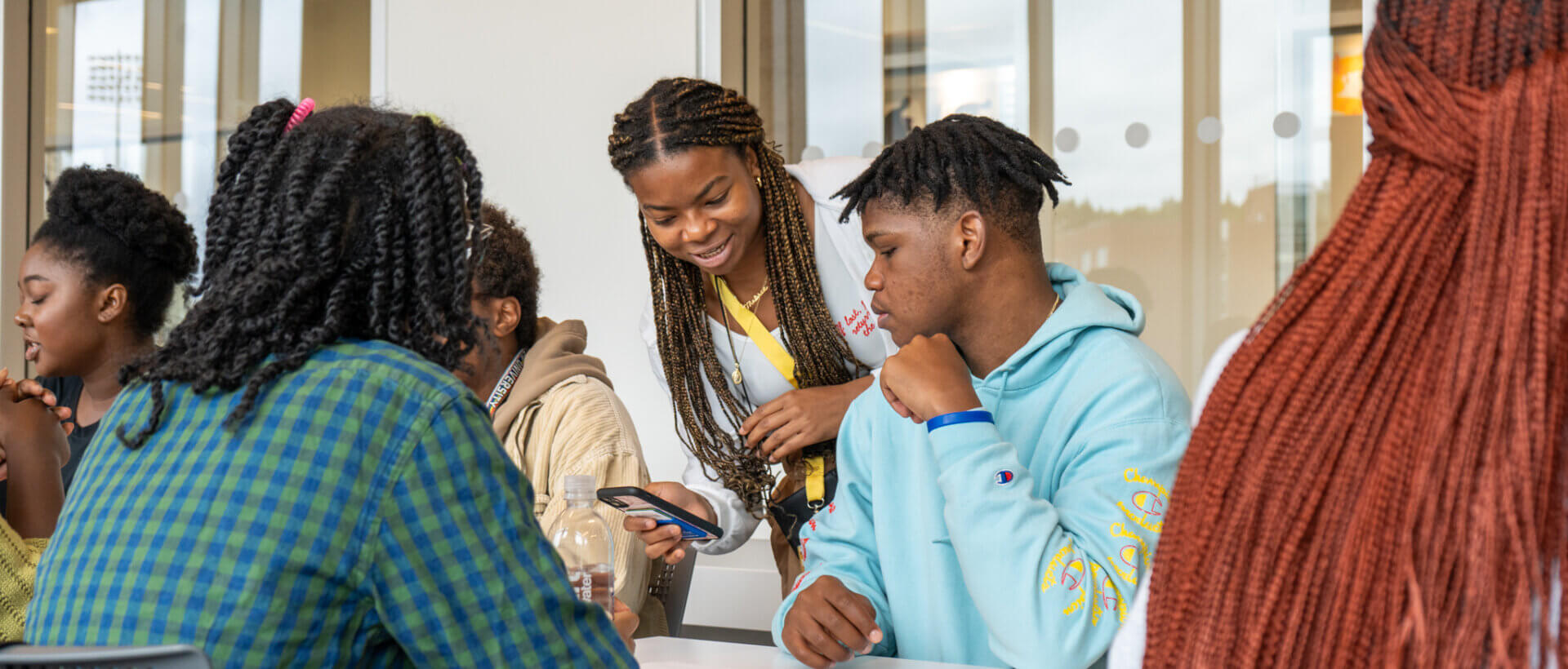 A group of young people sit at a table in a bright room, engaged and smiling. One person stands, showing something on a phone to the others. Friendly atmosphere.