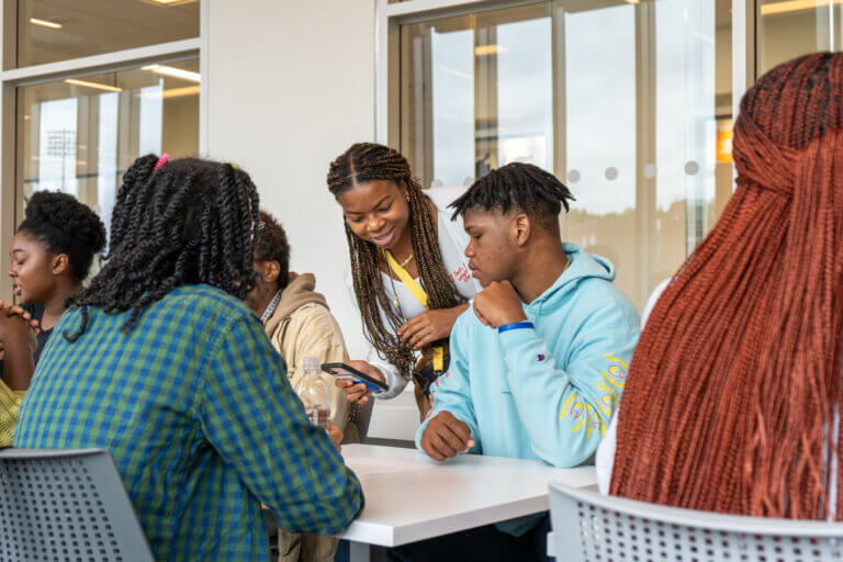 A group of young people sit at a table in a bright room, engaged and smiling. One person stands, showing something on a phone to the others. Friendly atmosphere.