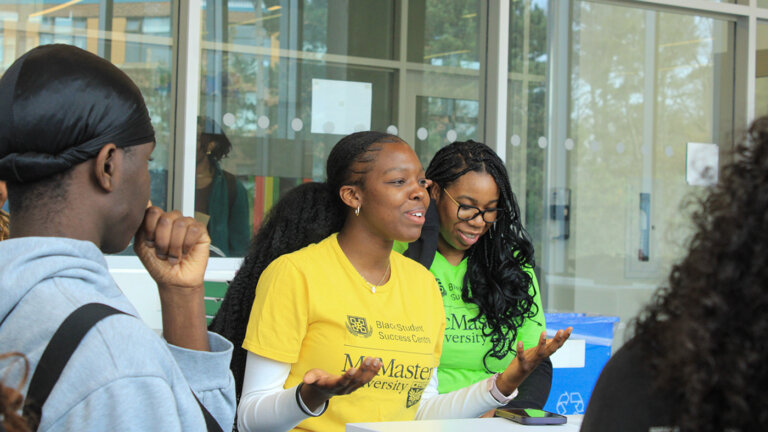 Two people in colorful shirts, smiling and engaged in a lively conversation indoors, with others nearby. The atmosphere is positive and collaborative.