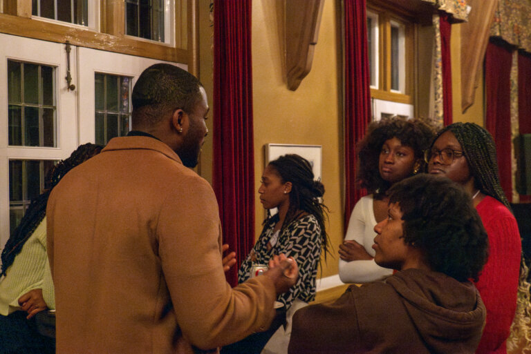 A group of people stand in a dimly lit room, engaged in conversation. The atmosphere is warm and focused, with rich red curtains in the background and large windows.