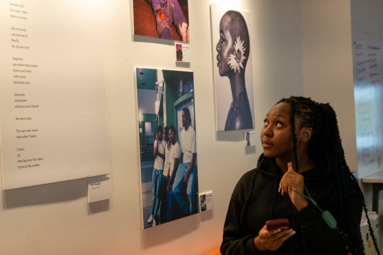 Young woman in a black hoodie looks thoughtfully at a wall display of photographs and text, holding a phone. The scene conveys contemplation.
