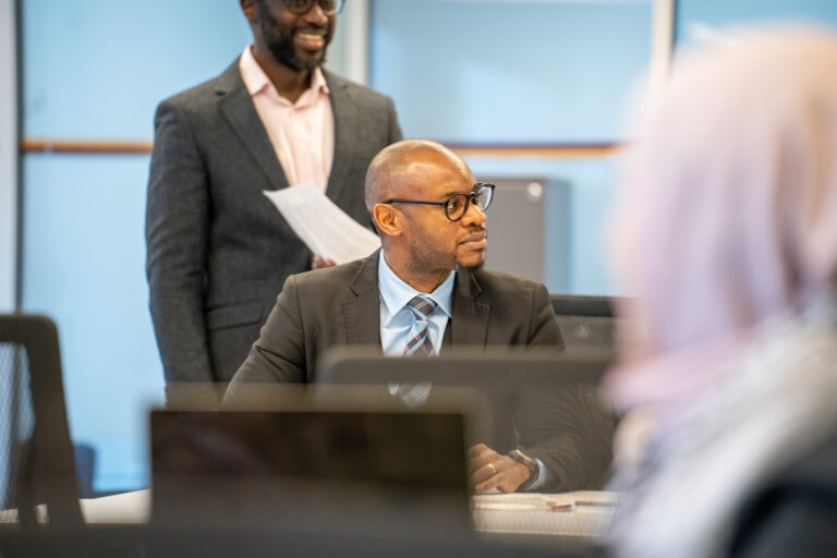 A professional man in a suit and glasses sits at a desk, attentively looking sideways. A smiling colleague stands holding papers. Office setting, collaborative tone.