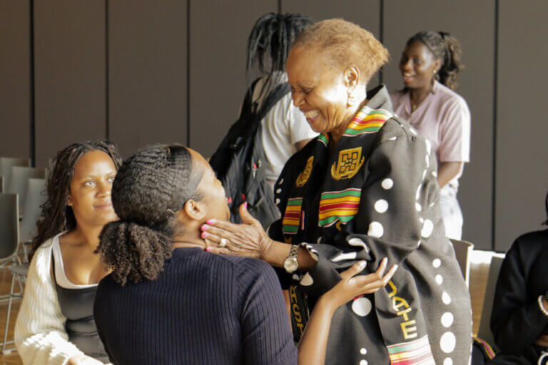 A woman wearing a colorful kente stole joyfully embraces a seated woman in a warm, congratulatory gesture. Onlookers smile, creating a celebratory atmosphere.