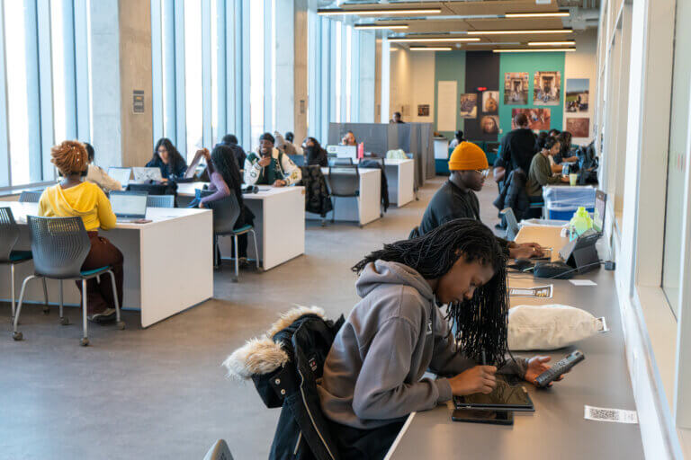 A well-lit study area with large windows, where diverse students work at desks with laptops and tablets. The atmosphere is focused and collaborative.
