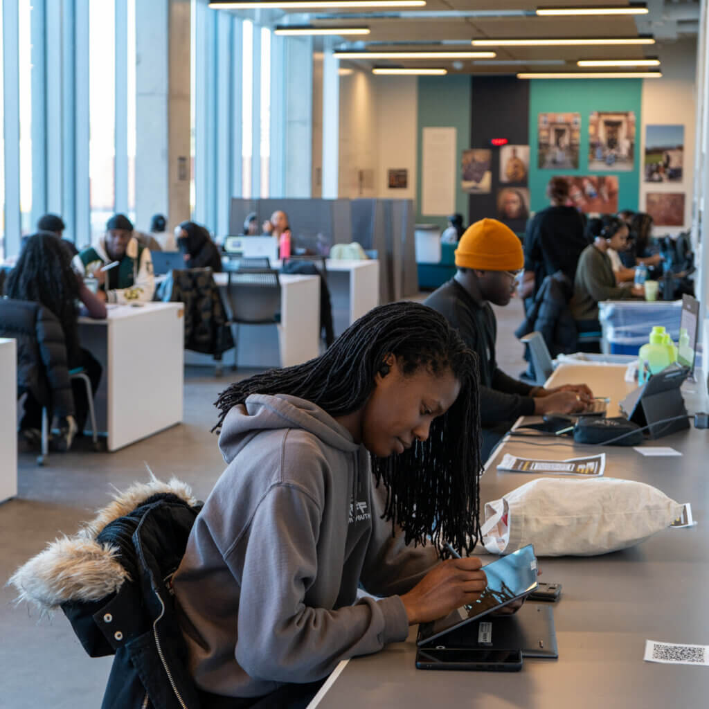 Students study in a modern library, sitting at long tables with laptops and tablets. The setting is bright and focused, with large windows and academic decor.
