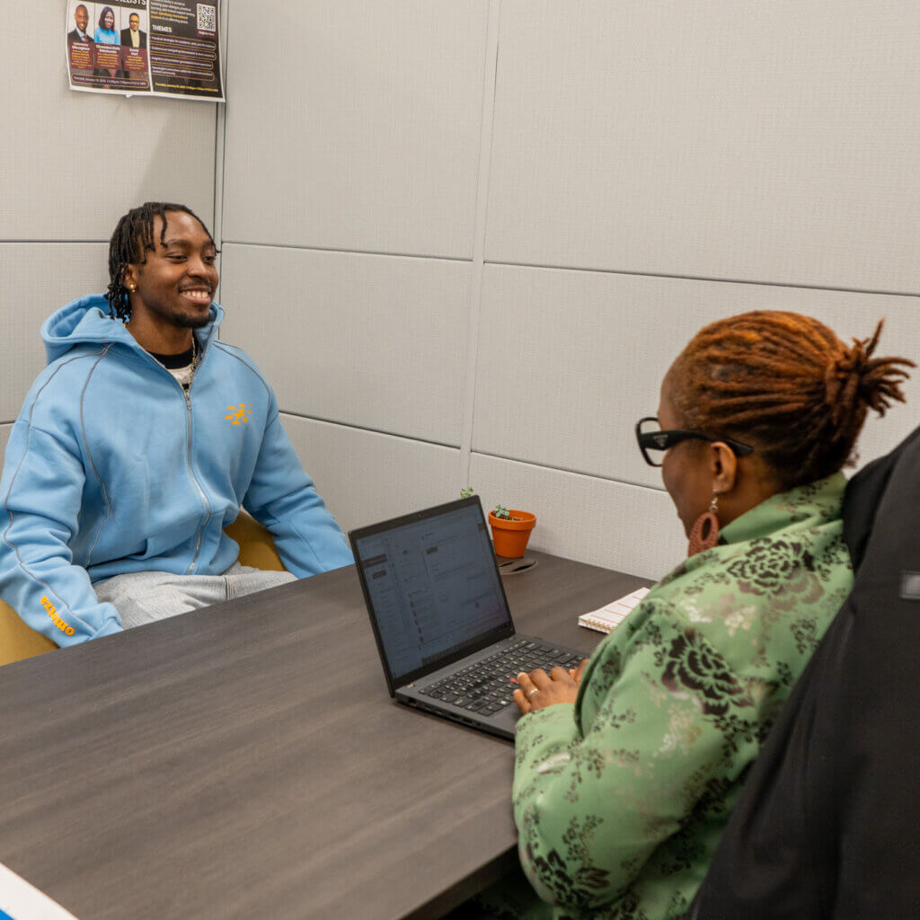 A man in a blue hoodie smiles while sitting across a table from a woman with glasses using a laptop. Both are in an office setting.