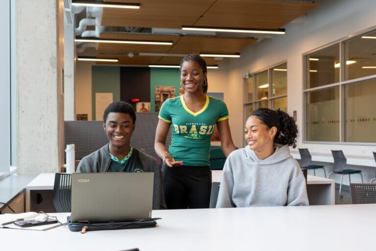 A woman stands smiling behind two seated people who are looking at a laptop in a modern workspace. The mood is collaborative and cheerful.