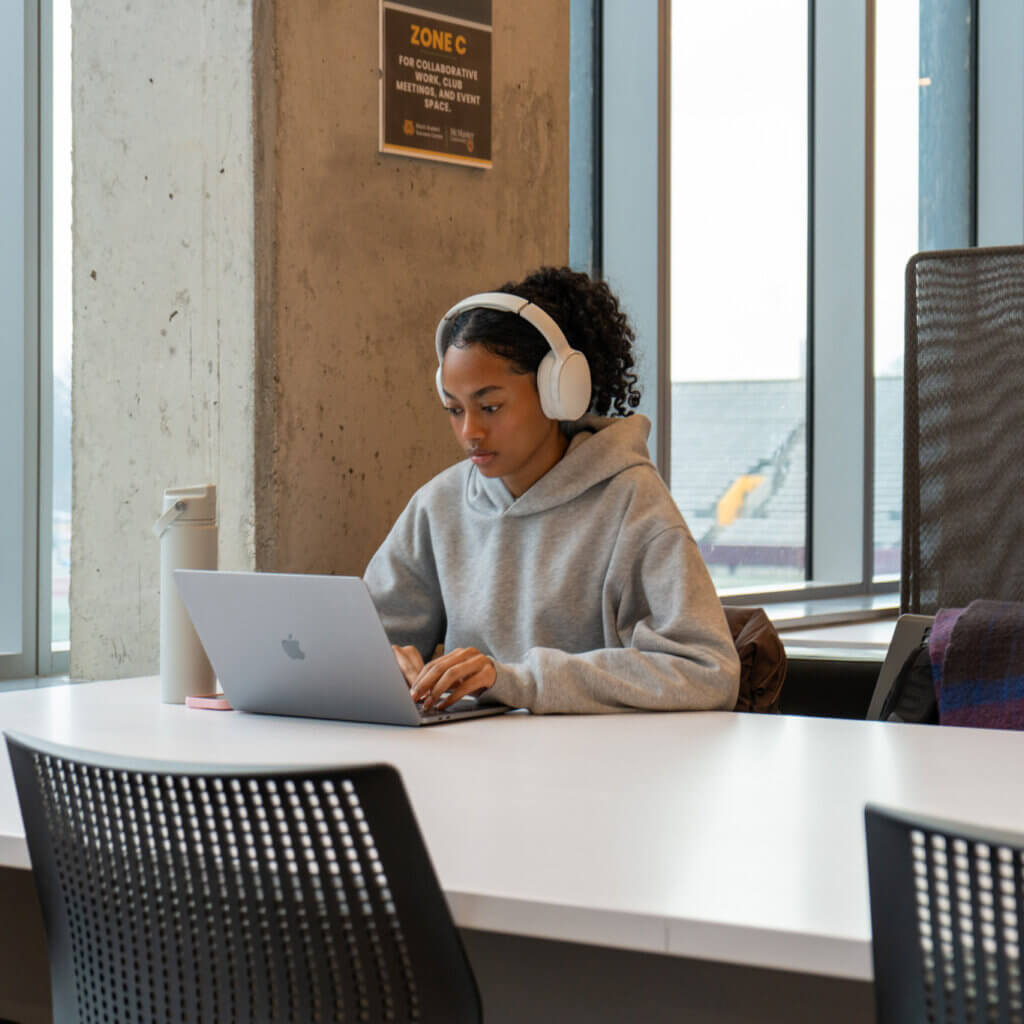 A person sits by a large window at a white table, wearing headphones and typing on a laptop. They are focused and wearing a gray hoodie.