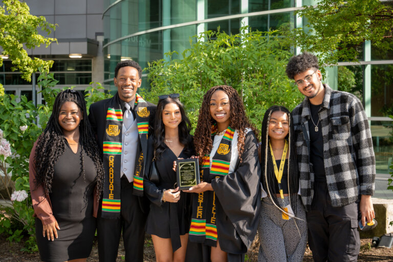 Six smiling graduates stand together outside, wearing colorful Kente stoles. One holds a plaque. The mood is celebratory and proud.