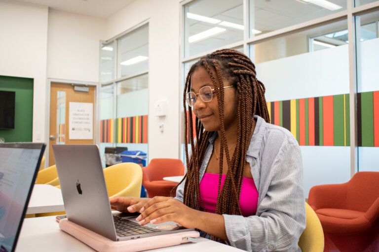 A woman with braided hair and glasses works on a laptop in a brightly colored, modern office. She appears focused and engaged, sitting at a table.