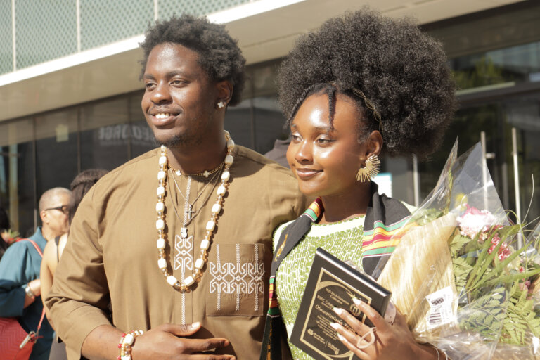 A smiling graduate wearing a green and black outfit holds a diploma and flowers, standing beside a man in a brown traditional shirt and shell necklace.