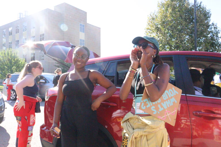 Two women stand smiling by a red car on a sunny day. One wears sunglasses and a black dress; the other holds a megaphone and a sign. People and a building are in the background.