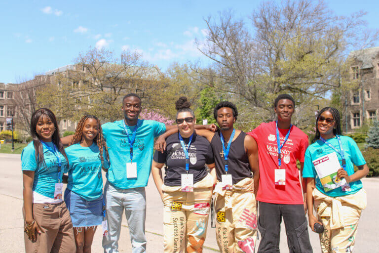 A group of seven people, some wearing blue and red McMaster University shirts, smile outdoors on a sunny day. They have name tags and embrace casually.