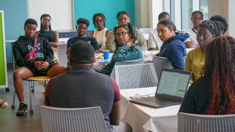 A group of individuals sits attentively around tables in a room, listening to a speaker. The atmosphere is engaged, with a laptop open in the foreground.