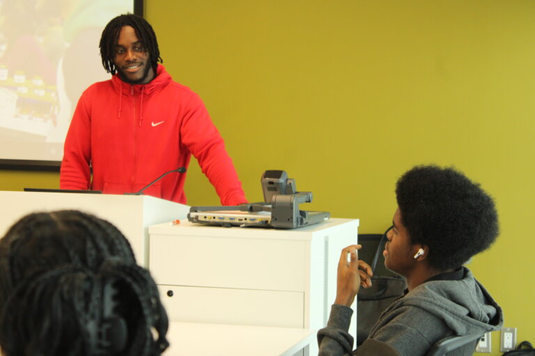 A man in a red hoodie speaks at a podium, smiling at a student with headphones in a classroom setting. The tone is casual and engaging.