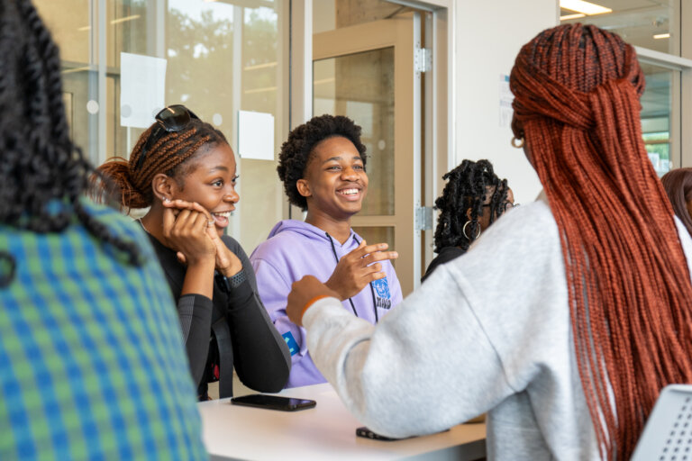 A group of students sits at a table, engaged in lively conversation. They are smiling and laughing, creating a joyful and relaxed atmosphere in a classroom setting.