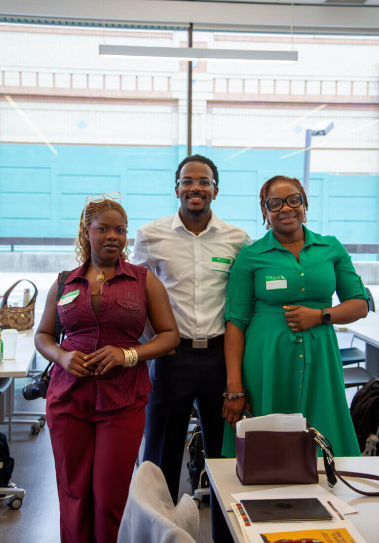Three people smiling in an office setting, standing side by side. They're wearing smart-casual attire with name badges, conveying a friendly mood.