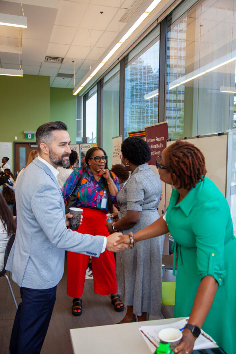 A man in a light blue blazer shakes hands with a woman in a green dress at a networking event. Two women in the background, one in colorful attire, are engaged in conversation. The atmosphere is professional and friendly.